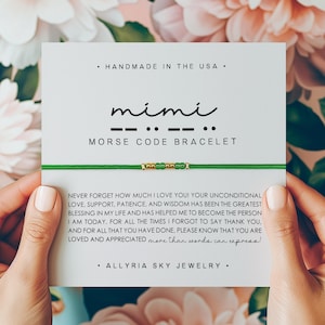 May include: A green string bracelet with gold beads that spells out "mimi" in Morse code. The bracelet is on a white card with the text "Handmade in the USA" and "Morse Code Bracelet". The card also includes a message about the importance of love, support, patience, and wisdom. The card is signed "Allryia Sky Jewelry."
