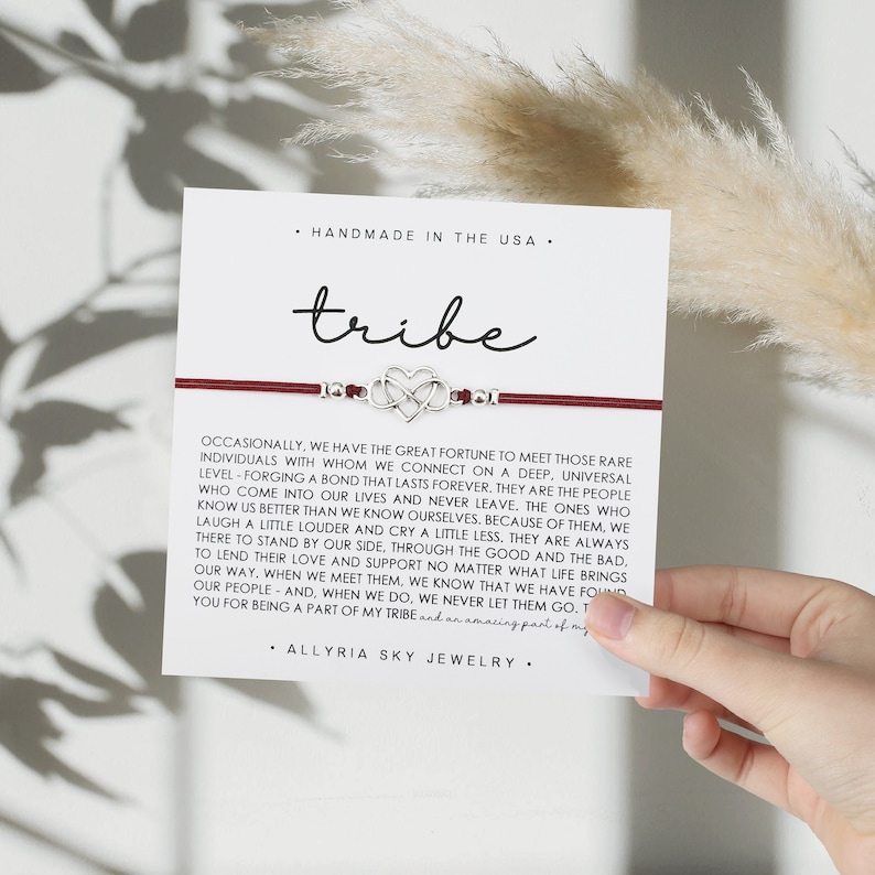 May include: A red string bracelet with a silver charm of two intertwined hearts. The bracelet is on a white card with the text "tribe" and a quote about the importance of finding your tribe.