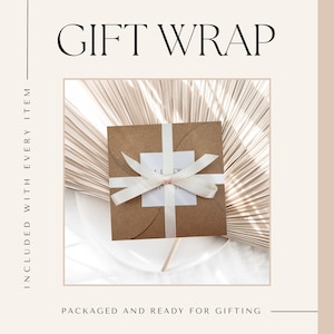 May include: A brown paper gift box with a white satin ribbon tied in a bow. The box is sitting on a white surface with a light brown palm leaf background.