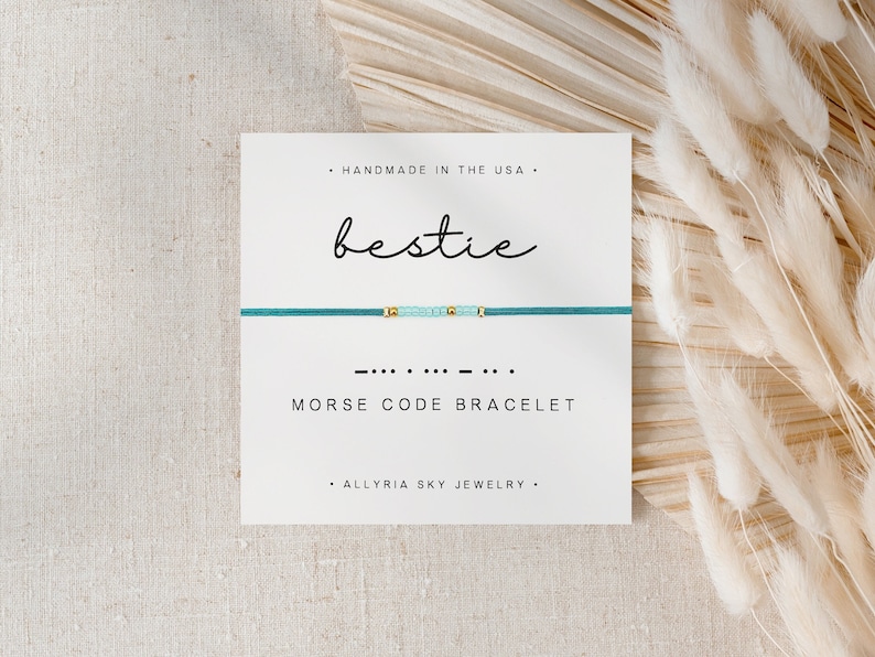 May include: A teal blue friendship bracelet with gold beads spelling out "bestie" in Morse code. The bracelet is displayed on a white card with the text "Handmade in the USA", "Bestie", "Morse Code Bracelet", and "Allyria Sky Jewelry".