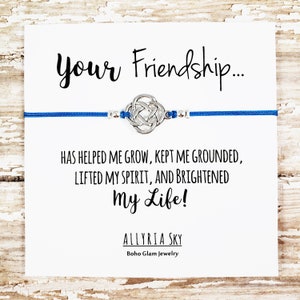 Puede incluir: Un brazalete de amistad azul con un dije de nudo plateado y el texto "Your Friendship... Has helped me grow, kept me grounded, lifted my spirit, and brightened my life! Allyria Sky Boho Glam Jewelry".