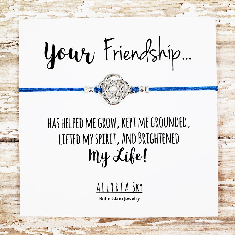 Puede incluir: Un brazalete de amistad azul con un dije de nudo plateado y el texto "Your Friendship... Has helped me grow, kept me grounded, lifted my spirit, and brightened my life! Allyria Sky Boho Glam Jewelry".