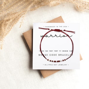 May include: A red string bracelet with silver beads spelling out "warrior" in Morse code. The bracelet is displayed on a white card with the text "Handmade in the USA", "Morse Code Bracelet", and "Allyria Sky Jewelry".