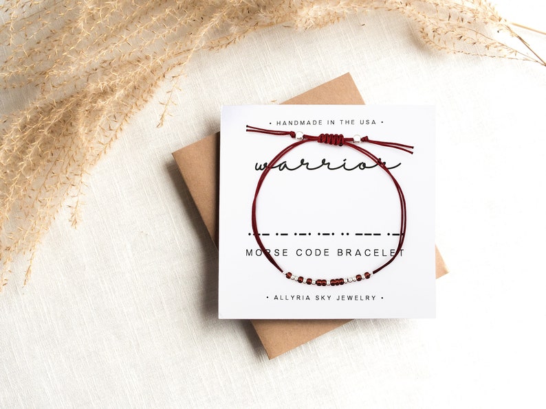 May include: A red string bracelet with silver beads spelling out "warrior" in Morse code. The bracelet is displayed on a white card with the text "Handmade in the USA", "Morse Code Bracelet", and "Allyria Sky Jewelry".