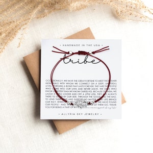 May include: A burgundy friendship bracelet with silver beads and the word "tribe" printed on a white card. The card also includes a quote about the importance of friendship.