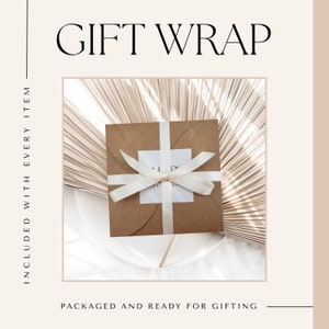 May include: A brown paper gift box with a white satin ribbon tied in a bow. The box is sitting on a white surface with a light brown palm leaf background.