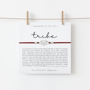 May include: A red string bracelet with a silver infinity symbol charm. The bracelet is on a white card with the word "tribe" written in black cursive. The card also includes a quote about the importance of finding your tribe.