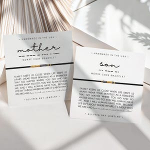 May include: Two white cards with black text and a black string bracelet with gold and gray beads. The text on the cards reads "Handmade in the USA. Mother ... Morse Code Bracelet" and "Handmade in the USA. Son ... Morse Code Bracelet". The text on the cards also includes a message about family and love.