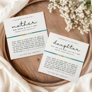 Peut inclure: Deux cartes blanches avec des bracelets bleus et dorés. Les cartes portent les mots "mère" et "fille" écrits en noir. Le texte sous les mots dit "Bracelet en code Morse". Les cartes contiennent également un message sur la famille et l'amour.