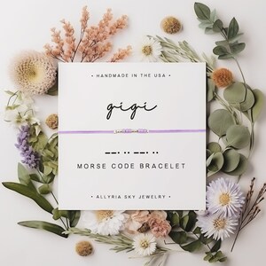 May include: A purple and silver bracelet with the word "gigi" written in cursive on a white card. The card also says "Morse Code Bracelet" and "Handmade in the USA".