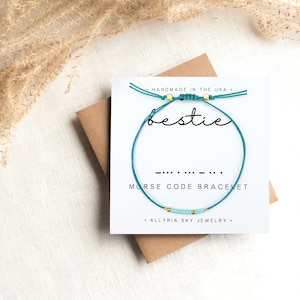 May include: Teal blue friendship bracelet with gold beads and the word "bestie" written on a white card. The bracelet is a morse code bracelet with the morse code for "bestie" written below the word. The card also says "Handmade in the USA" and "Allryia Sky Jewelry".