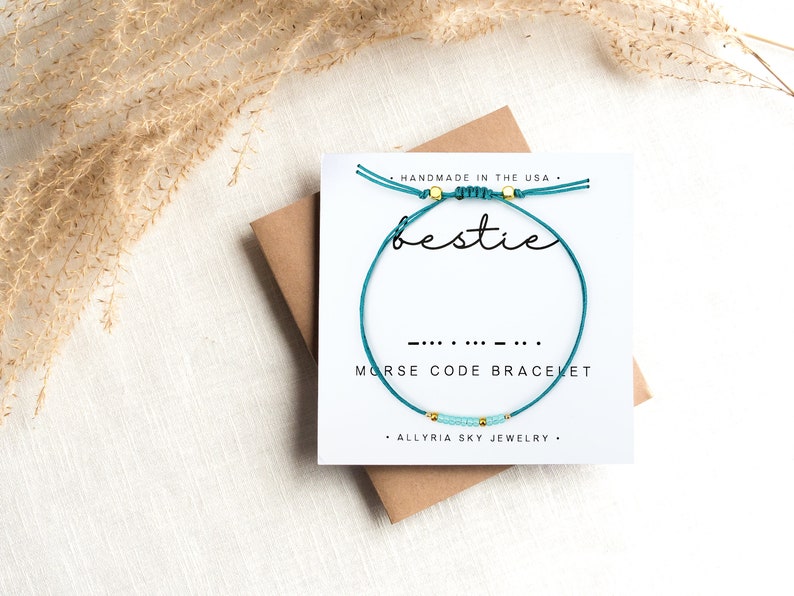 May include: Teal blue friendship bracelet with gold beads and the word "bestie" written on a white card. The bracelet is a morse code bracelet with the morse code for "bestie" written below the word. The card also says "Handmade in the USA" and "Allryia Sky Jewelry".