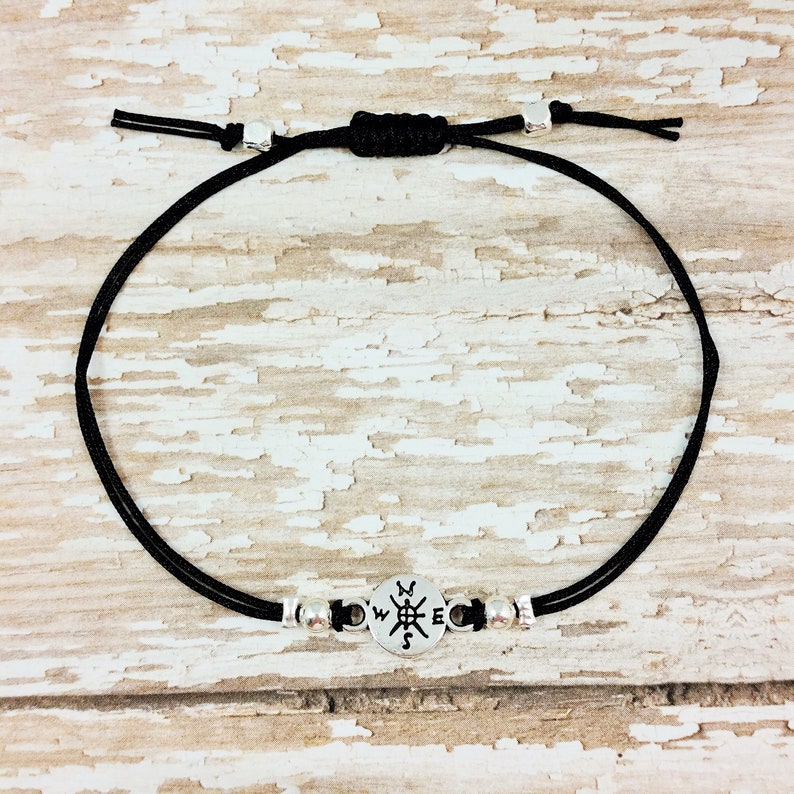 May include: A black cord bracelet with silver beads and a compass charm. The compass charm has the letters N, S, E, and W engraved on it.
