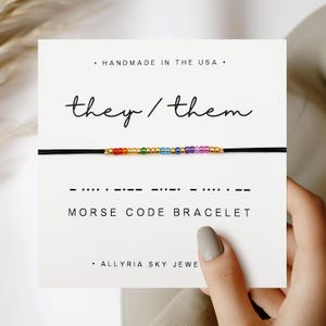 Peut inclure: Un bracelet en cordon noir avec des perles arc-en-ciel qui épellent "they/them" en code Morse. Le bracelet est présenté sur une carte blanche avec le texte "HANDMADE IN THE USA", "they/them", "MORSE CODE BRACELET", et "ALLYRIA SKY JEWE".