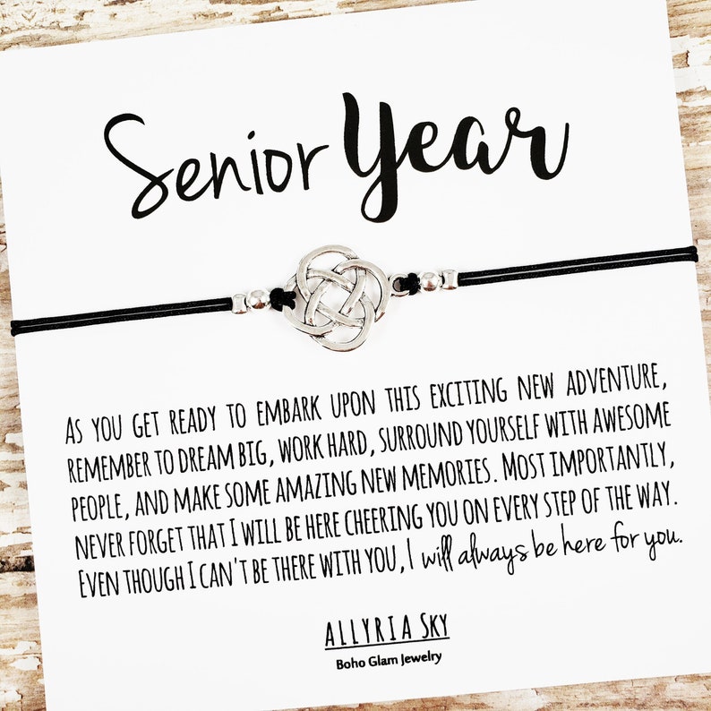 May include: A black string bracelet with a silver Celtic knot charm. The bracelet has a card attached with the text "Senior Year" and a message about embarking on a new adventure, working hard, and making new memories. The card is signed "Allyria Sky, Boho Glam Jewelry."