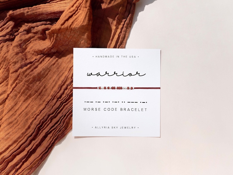 May include: A red string bracelet with silver beads spelling out "warrior" in Morse code. The bracelet is displayed on a white card with the words "Handmade in the USA", "Morse Code Bracelet", and "Allryia Sky Jewelry".