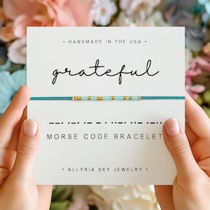 May include: A teal blue string bracelet with gold and light blue beads. The bracelet spells out the word "grateful" in Morse code. The card reads "Handmade in the USA. Grateful. --. ... ... Morse Code Bracelet. Allyria Sky Jewelry."