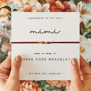 May include: A red string bracelet with gold beads spelling out the name "mimi" in Morse code. The bracelet is displayed on a white card with the text "HANDMADE IN THE USA", "mimi", "--:--:", "MORSE CODE BRACELET", and "ALLYRIA SKY JEWELRY".