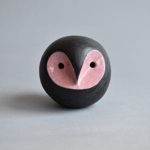 May include: A spherical ceramic sculpture shaped like an owl with a matte black finish and a glossy, heart-shaped light pink face. The owl features two small, dark, circular indentations for eyes centered within the pink facial disc.