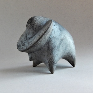 Può includere: Una scultura in ceramica grigia di un animale astratto con una grande testa arrotondata e tre gambe. La scultura ha una superficie testurizzata e una finitura usurata.