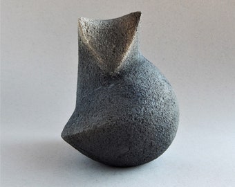 May include: A small, abstract ceramic sculpture with a rough, textured surface. The sculpture is a dark gray color with a lighter gray area on the top. It is shaped like a rounded, asymmetrical form.