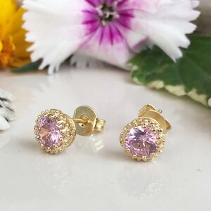 Puede incluir: Un par de pendientes de bot&oacute;n de oro con una piedra preciosa rosa en el centro de cada pendiente. Los pendientes tienen un dise&ntilde;o delicado y ornamentado alrededor de la piedra preciosa.