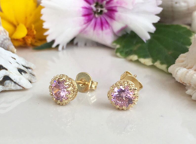 Puede incluir: Un par de pendientes de bot&oacute;n de oro con una piedra preciosa rosa en el centro de cada pendiente. Los pendientes tienen un dise&ntilde;o delicado y ornamentado alrededor de la piedra preciosa.