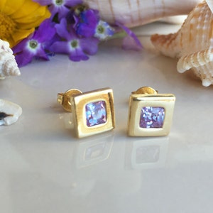 May include: A pair of gold square stud earrings with a light purple square gemstone in the center of each earring.
