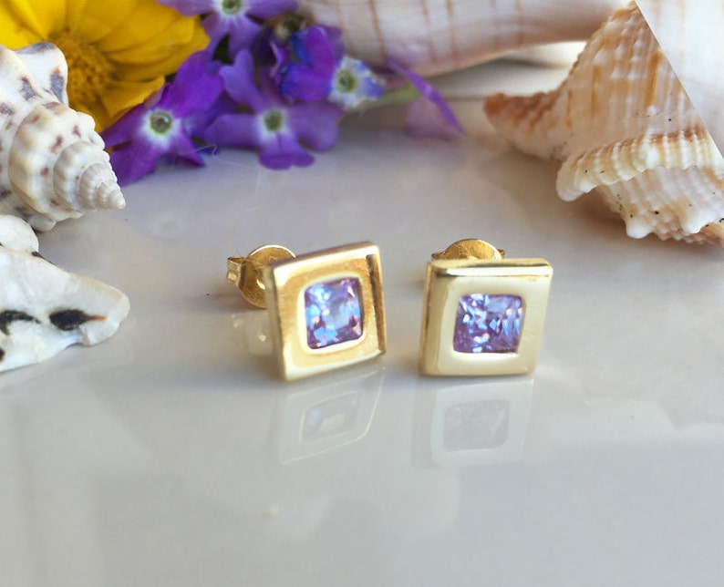 May include: A pair of gold square stud earrings with a light purple square gemstone in the center of each earring.