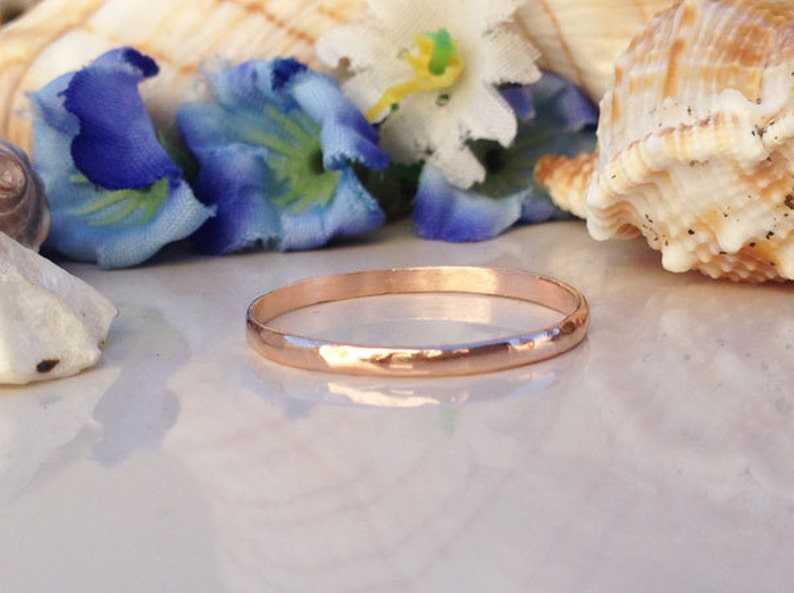 May include: A thin rose gold band ring with a hammered finish. The ring is displayed on a white surface with seashells and flowers.