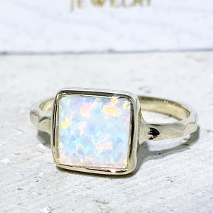 May include: A gold ring with a square-shaped opal gemstone. The opal is iridescent with shades of blue, pink, and white.
