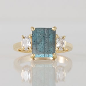 Könnte beinhalten: Ein goldener Ring mit einem großen, rechteckigen, blaugrünen Edelstein und zwei kleineren, rechteckigen, klaren Edelsteinen auf jeder Seite.