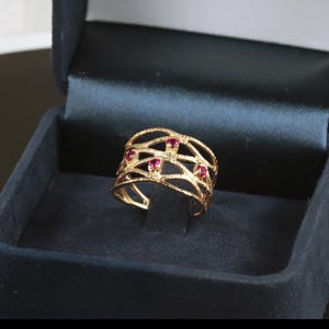 以下が含まれることがあります： 繊細な編み込みのデザインと、小さな赤い宝石が5つ付いた金の指輪です。