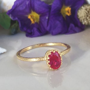 May include: A gold ring with a bright red oval gemstone set in a decorative gold setting. The ring is on a white surface.