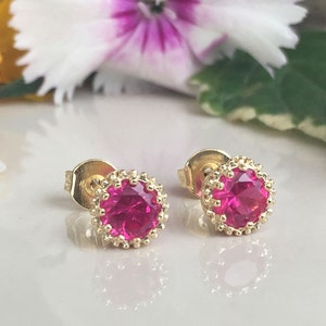 May include: A pair of gold stud earrings with a pink gemstone in the center of each earring. The earrings have a decorative gold border around the gemstone.