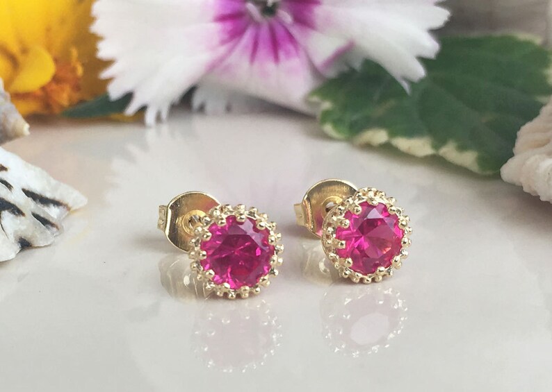 May include: A pair of gold stud earrings with a pink gemstone in the center of each earring. The earrings have a decorative gold border around the gemstone.