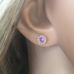 Puede incluir: Un par de pendientes de oro con una peque&ntilde;a piedra preciosa redonda de color p&uacute;rpura en el centro. La piedra preciosa est&aacute; rodeada por un halo de piedras doradas m&aacute;s peque&ntilde;as.