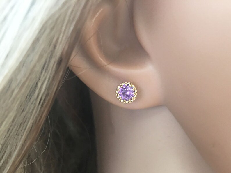 Puede incluir: Un par de pendientes de oro con una peque&ntilde;a piedra preciosa redonda de color p&uacute;rpura en el centro. La piedra preciosa est&aacute; rodeada por un halo de piedras doradas m&aacute;s peque&ntilde;as.