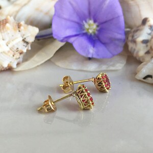 May include: A pair of gold earrings with red gemstone accents. The earrings are set against a background of seashells and a purple flower.