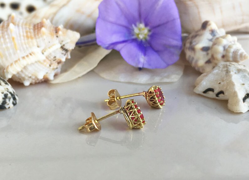 May include: A pair of gold earrings with red gemstone accents. The earrings are set against a background of seashells and a purple flower.