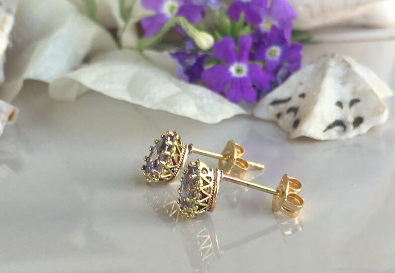 May include: A pair of gold stud earrings with a purple gemstone set in a crown-like design.