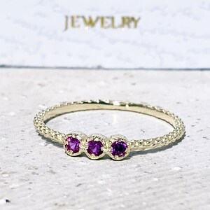 May include: A delicate gold band ring with three small purple gemstones set in a row. The band has a textured design.