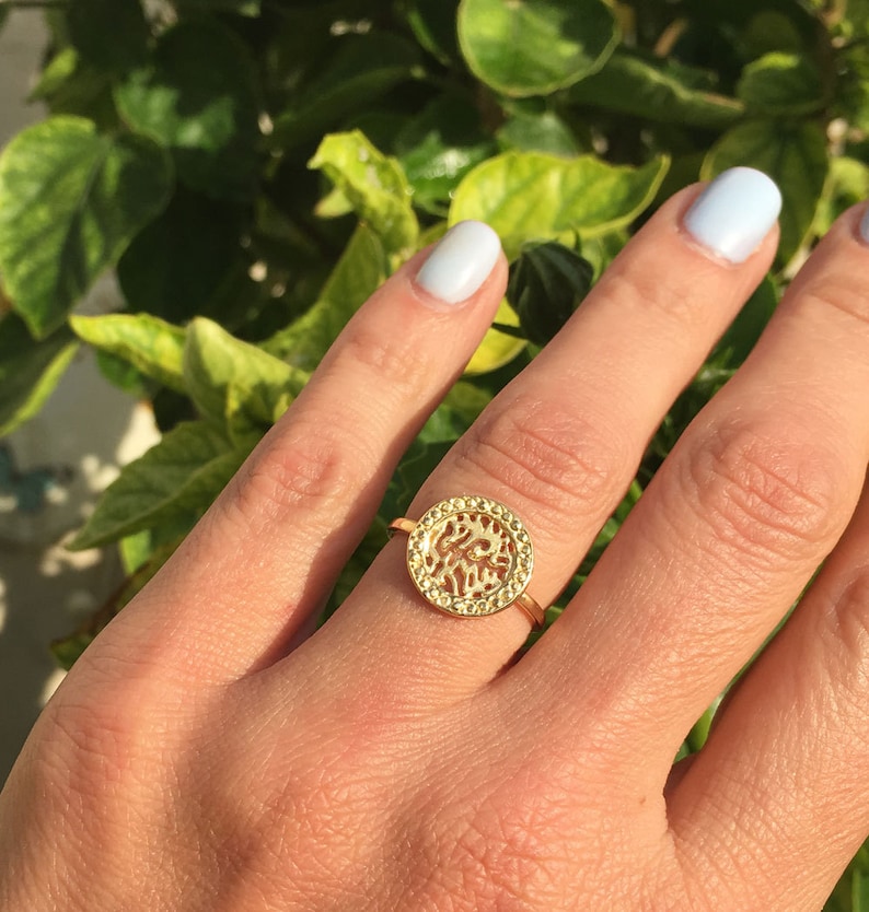 May include: A gold ring with a round, openwork design. The ring is on a finger and the background is a green leafy plant.