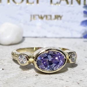 May include: A gold ring featuring a large oval purple gemstone and two smaller round clear gemstones. The ring is set in a plain band.