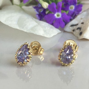 May include: A pair of gold stud earrings with a pear-shaped purple gemstone set in a gold bezel. The earrings have a delicate gold beaded border around the gemstone.