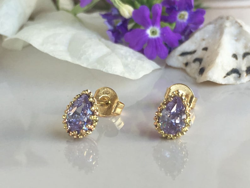 May include: A pair of gold stud earrings with a pear-shaped purple gemstone set in a gold bezel. The earrings have a delicate gold beaded border around the gemstone.