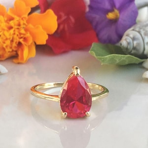 May include: A gold ring with a pear-shaped red gemstone. The ring is set against a white background with flowers and seashells.