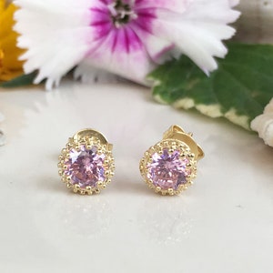 Puede incluir: Un par de pendientes de bot&oacute;n de oro con una piedra preciosa rosa en el centro de cada pendiente. Las piedras preciosas est&aacute;n rodeadas por un halo de oro.