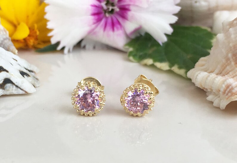 Puede incluir: Un par de pendientes de bot&oacute;n de oro con una piedra preciosa rosa en el centro de cada pendiente. Las piedras preciosas est&aacute;n rodeadas por un halo de oro.
