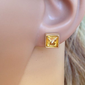 May include: A pair of gold square stud earrings with a yellow gemstone in the center.